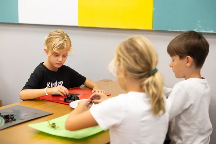 Drei Kinder arbeiten an einem Tisch mit dunkler Knete auf farbigen Unterlagen.