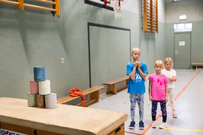 Drei Mädchen stehen in einer Turnhalle, im Vordergrund sind bunte Dosen auf einem Sprungtisch gestapelt.