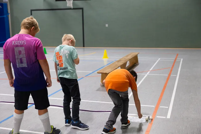 Drei Jungen in einer Sporthalle; einer im orangefarbenen T-Shirt bückt sich zu Gegenständen auf dem Boden.
