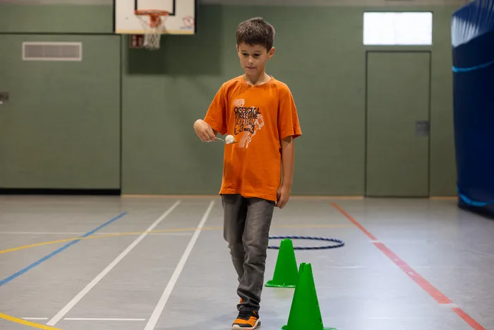 Ein Junge in einem orangefarbenen T-Shirt hält einen kleinen weißen Ball auf einem Stiel in einer Sporthalle.