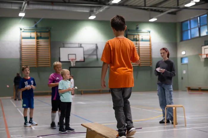 Ein Junge in einem orangefarbenen T-Shirt steht auf einer Holzbank in einer Turnhalle, während andere Kinder zusehen.