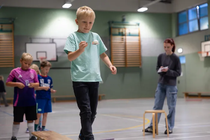 Ein Junge balanciert ein Ei auf einem Löffel, während er über einen Holzbalken in einer Turnhalle läuft.