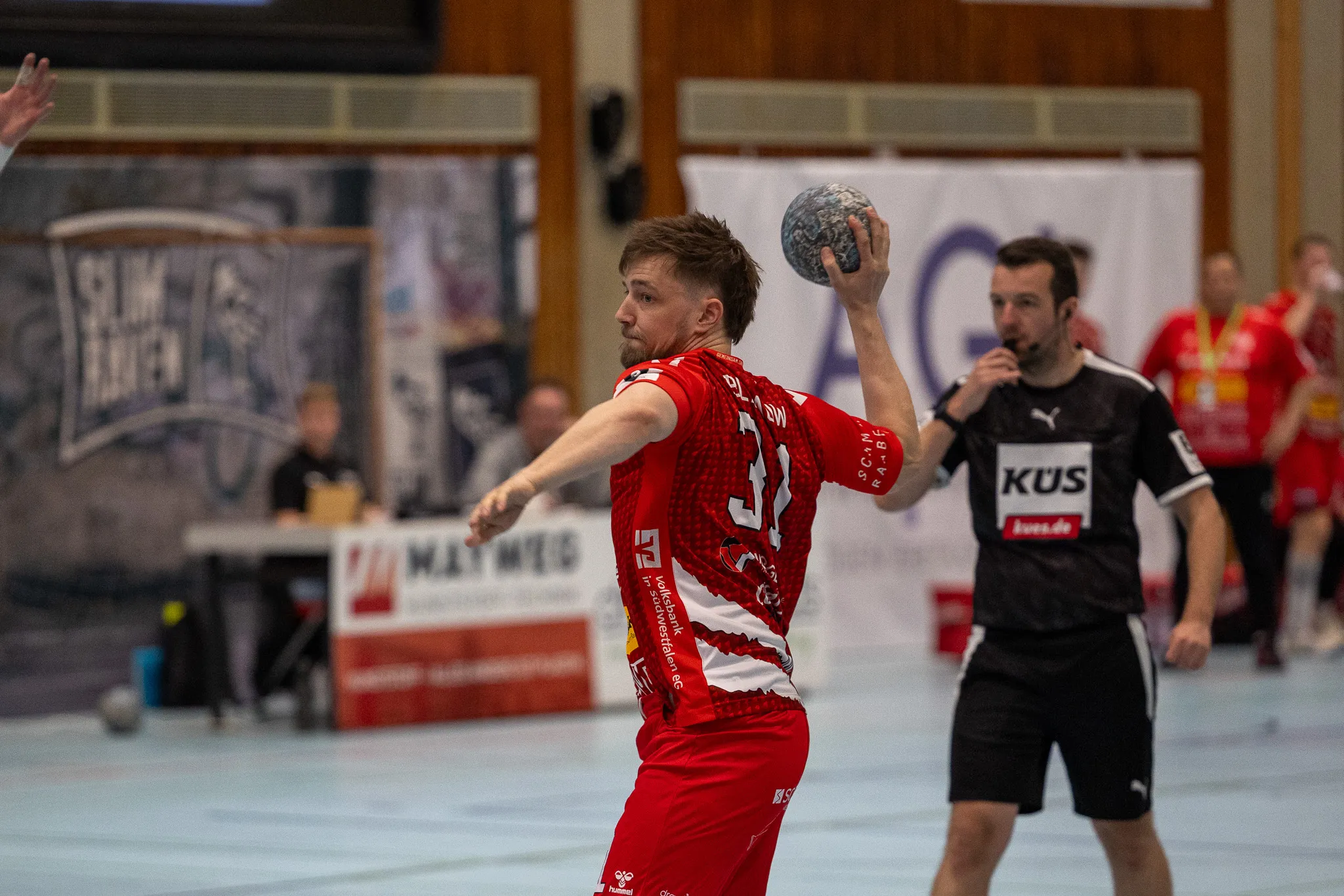 Handballspieler in rotem Trikot wirft den Ball während eines Spiels. Im Hintergrund weitere Spieler und Zuschauer in einer Sporthalle.