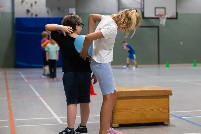 Zwei Kinder stehen in einer Turnhalle neben einer Holzkiste, eines hält einen blauen Luftballon.