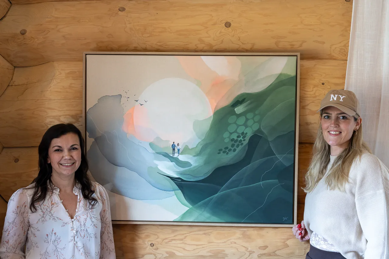 Zwei Frauen stehen vor einem großformatigen, abstrakten Gemälde einer Landschaft mit Bergen und einem orangefarbenen Himmel. Im Hintergrund ist eine Holzwand zu sehen.