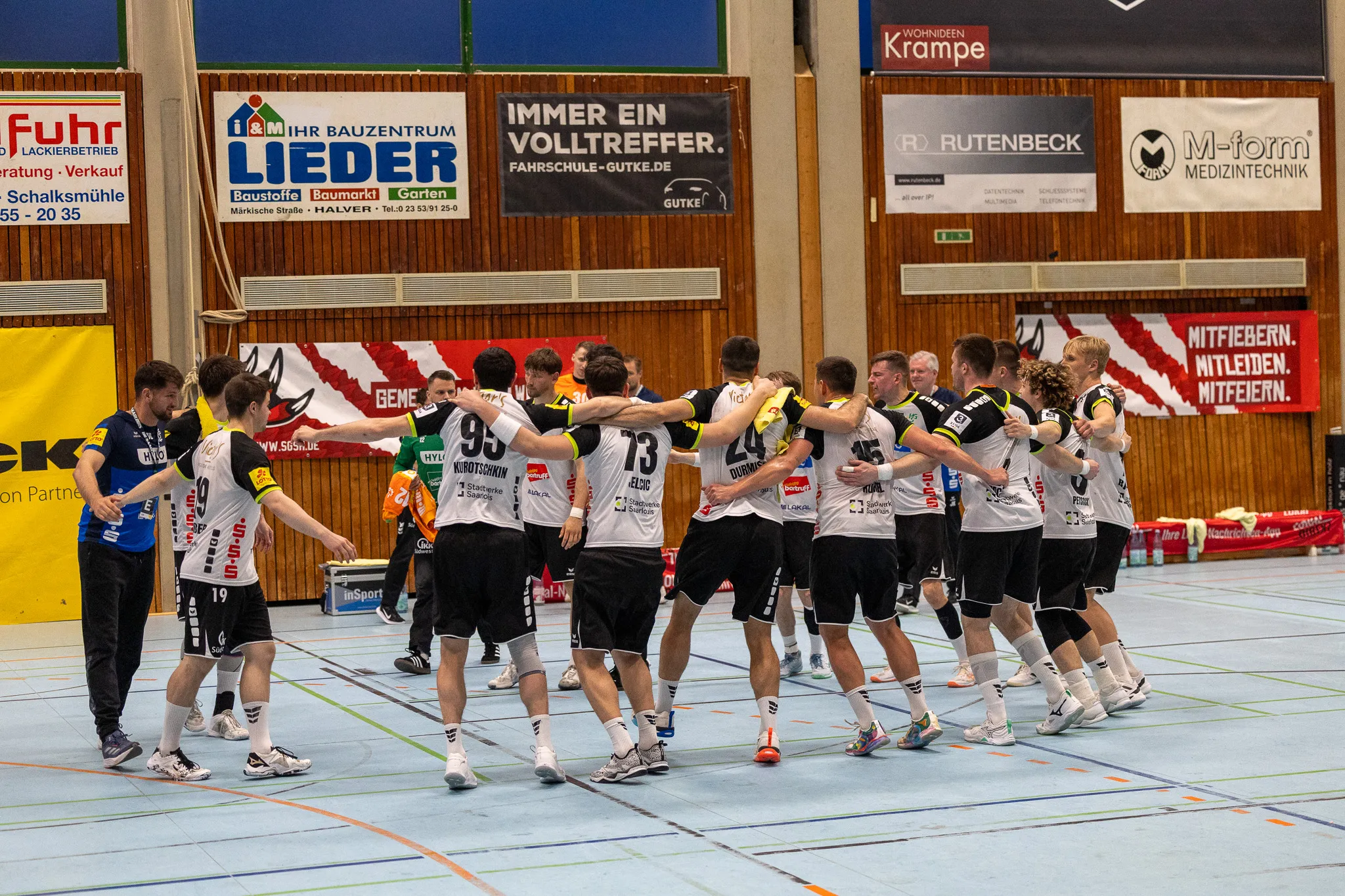 Handballspieler der SGSH Dragons umarmen sich nach einem Spiel in der Halle, während der Gegner im Hintergrund steht.