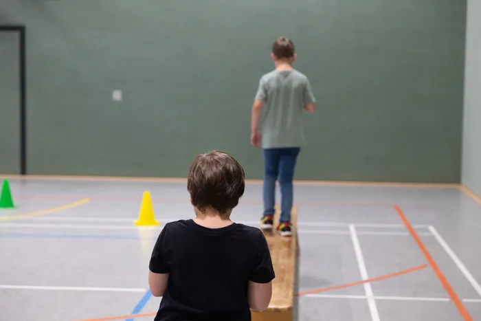 Ein Kind balanciert auf einem Holzbalken, während ein anderes Kind im Vordergrund zuschaut.