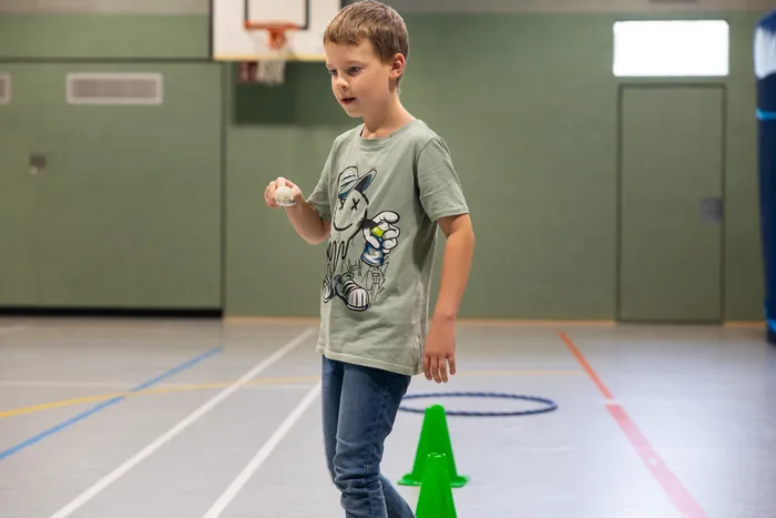 Ein Junge in einem grauen T-Shirt hält einen kleinen weißen Ball in der Hand in einer Turnhalle.