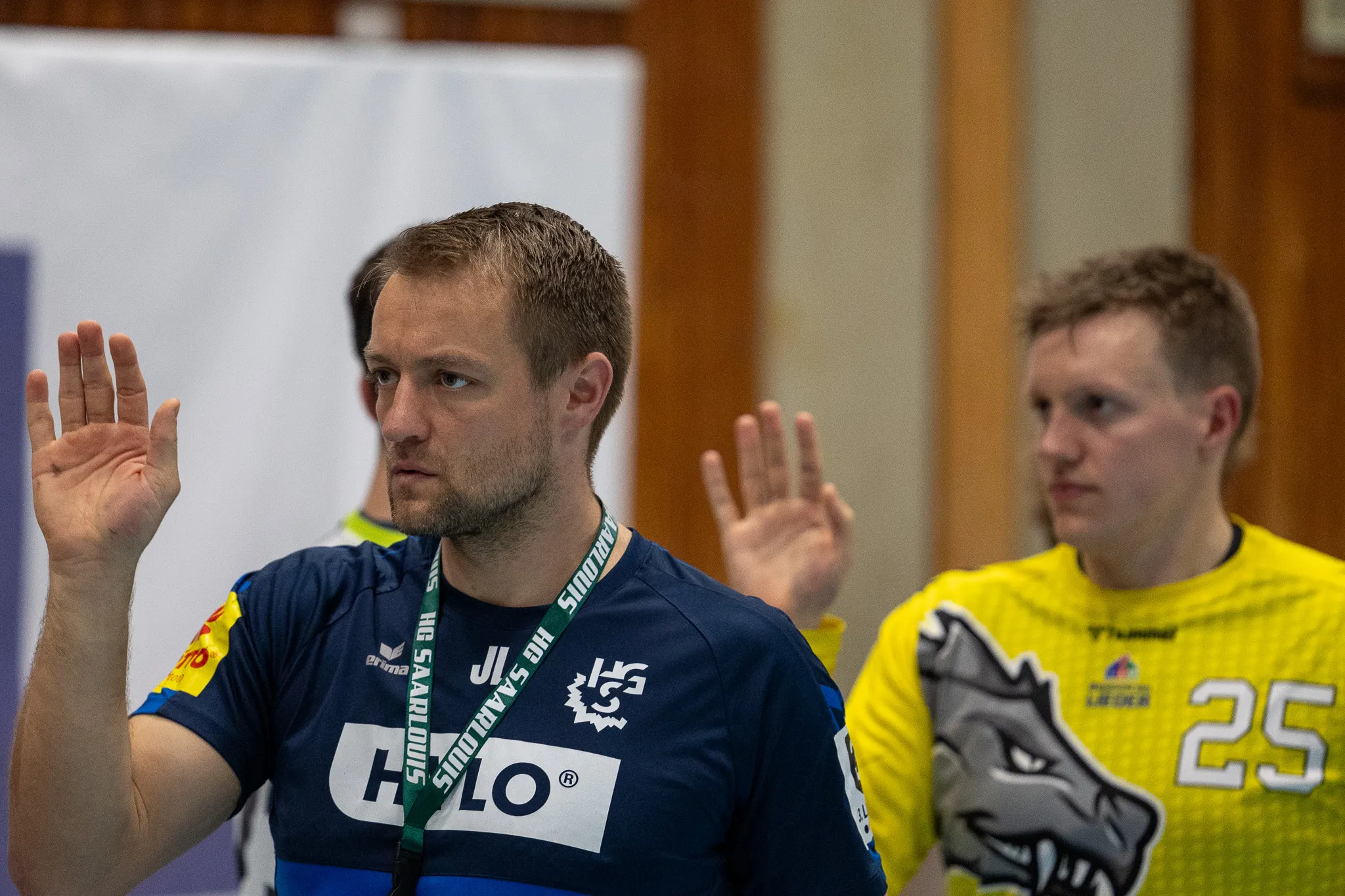 Handballspieler mit hochgehobenen Händen während des Spiels, im Hintergrund ein weiterer Spieler und die Tribüne.