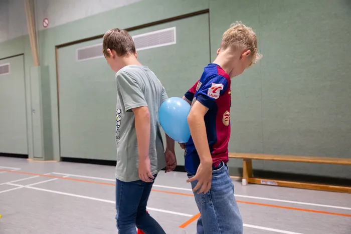Zwei Jungen halten einen blauen Luftballon zwischen ihren Rücken in einer Turnhalle.