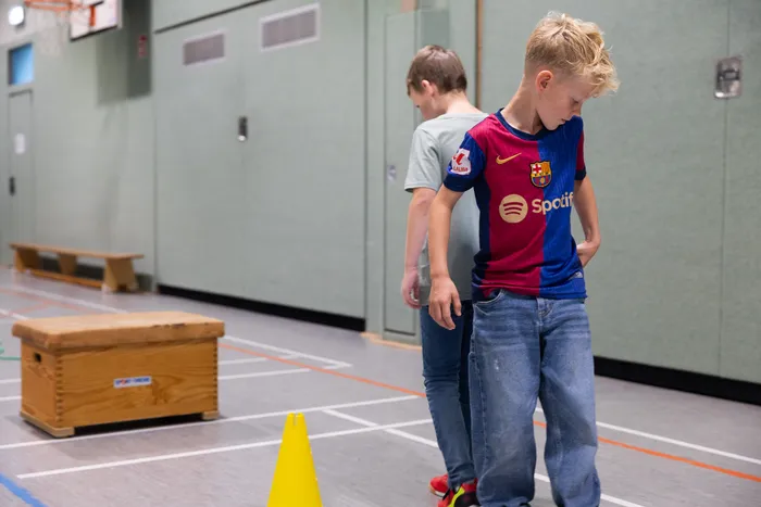 Zwei Jungen stehen in einer Turnhalle; einer trägt ein blau-rotes Fußballtrikot und blickt nach unten.