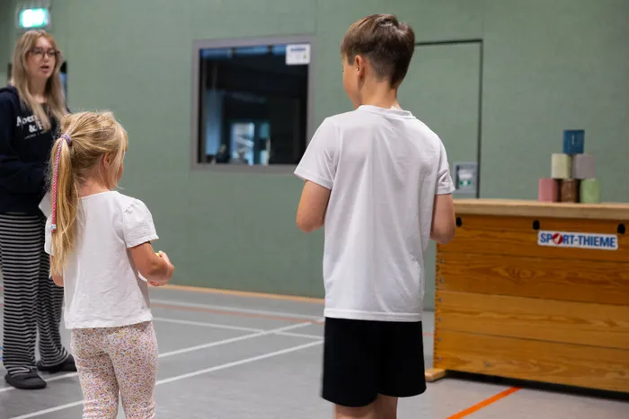 Ein Mädchen und ein Junge stehen in einer Turnhalle vor einem hölzernen Sportkasten.