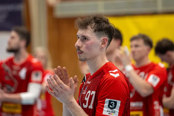 Handballspieler der SGSH Dragons mit der Trikotnummer 97 gestikuliert während eines Spiels. Im Hintergrund sind weitere Spieler in roten Trikots zu sehen.