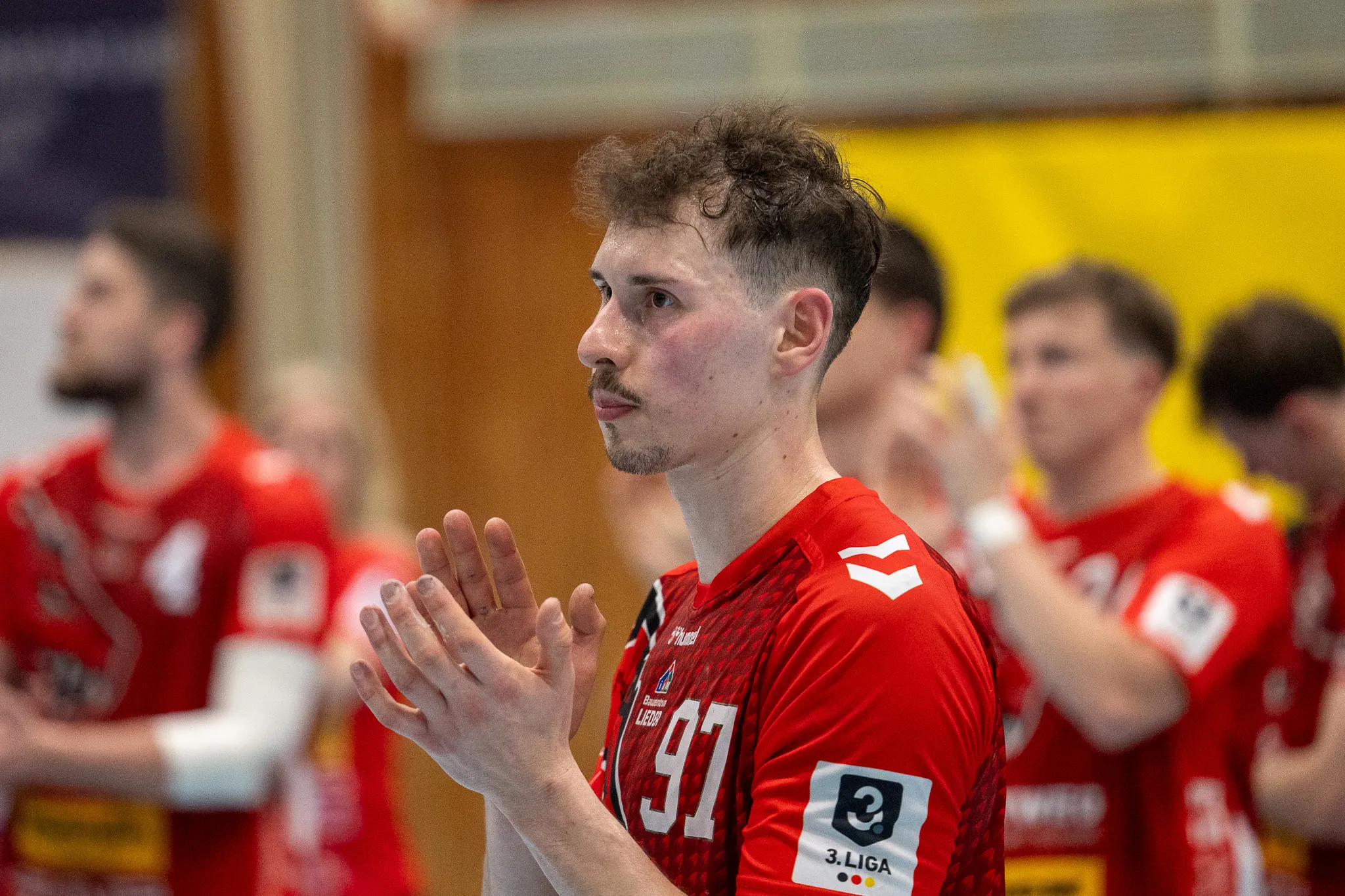 Handballspieler der SGSH Dragons mit der Trikotnummer 97 gestikuliert während eines Spiels. Im Hintergrund sind weitere Spieler in roten Trikots zu sehen.