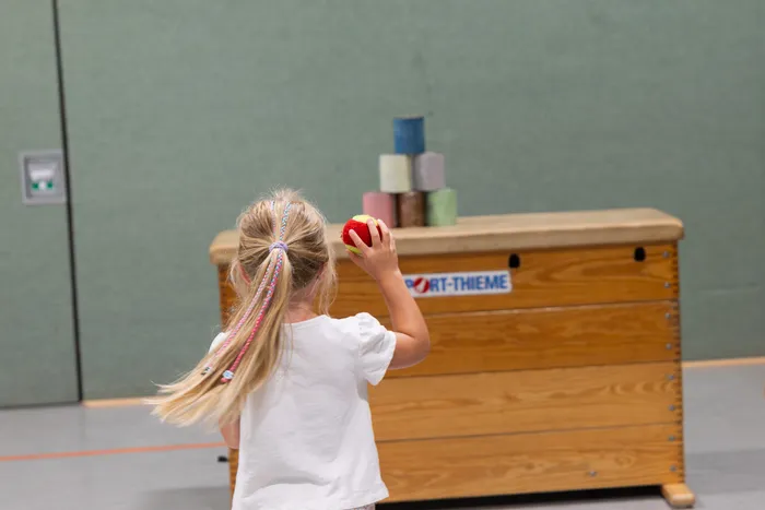Ein Mädchen mit blonden Zöpfen hält einen roten Ball hoch, um auf einen Stapel bunter Zylinder auf einem hölzernen Turnkasten zu zielen.