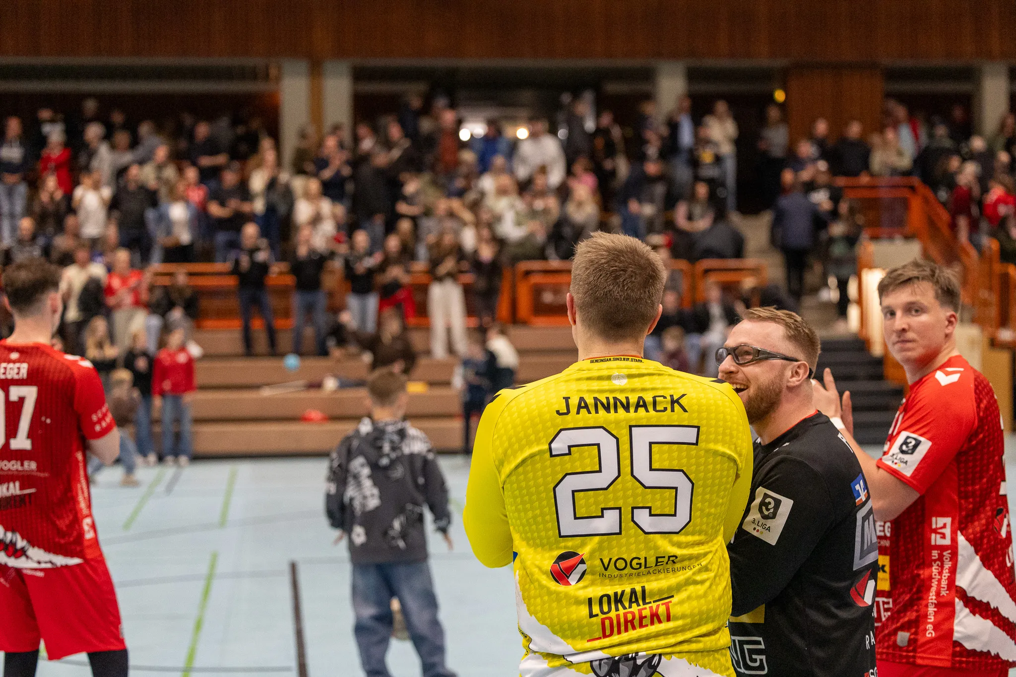 Handballspieler mit der Nummer 25 im gelben Trikot steht mit einem Schiedsrichter im Gespräch, während im Hintergrund Fans auf den Rängen sitzen.