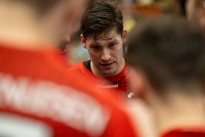 Handballspieler mit nassen Haaren und Blick zur Seite, trägt ein Trikot mit Hummel-Logo und steht inmitten anderer Spieler in roten Trikots.