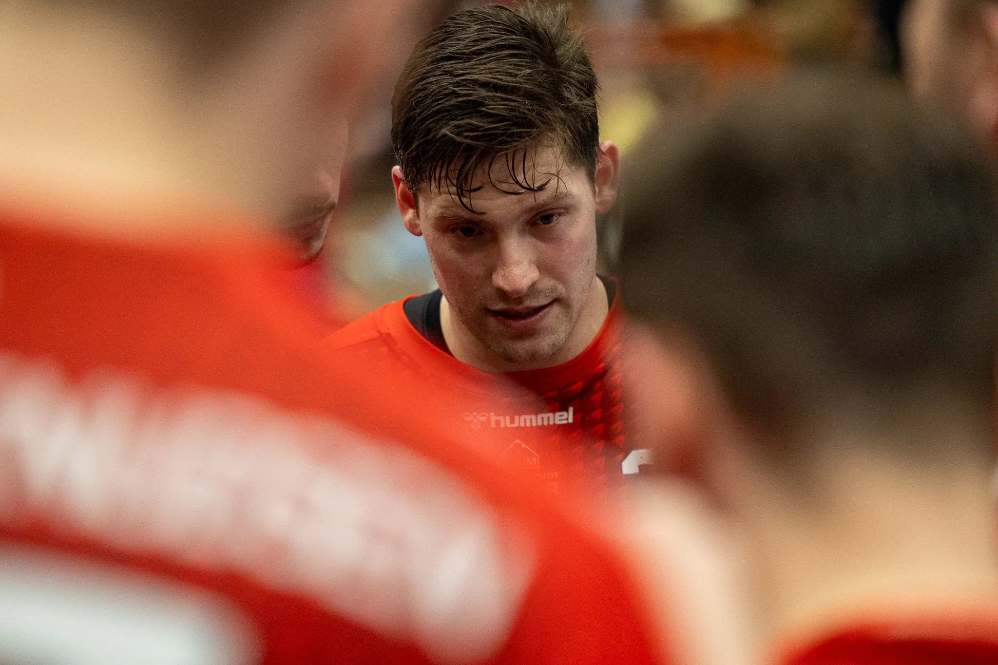 Handballspieler mit nassen Haaren und Blick zur Seite, trägt ein Trikot mit Hummel-Logo und steht inmitten anderer Spieler in roten Trikots.