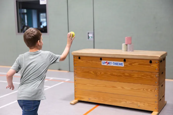 Ein Junge wirft einen gelben Ball auf eine hölzerne Sprungbox in einer Turnhalle.