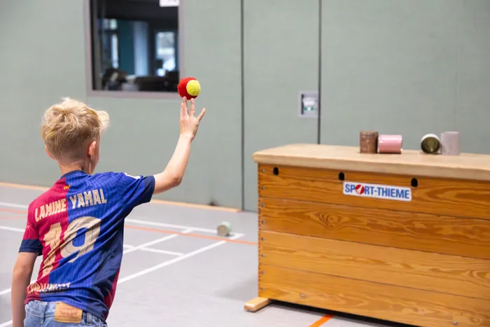 Ein Junge in einem blau-roten Fußballtrikot wirft einen rot-gelben Ball in einer Turnhalle nach oben.