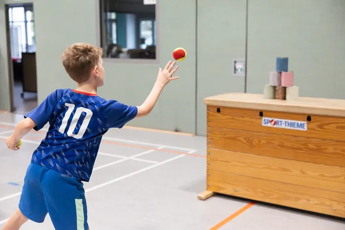 Ein Junge in blauem Sporttrikot mit der Nummer 10 wirft einen Ball auf einen Stapel kleiner Objekte auf einer hölzernen Turnbank.