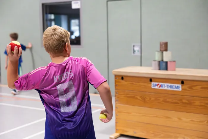 Ein Junge in einem pinkfarbenen Trikot bereitet in einer Sporthalle einen Wurf mit einem roten Ball vor.