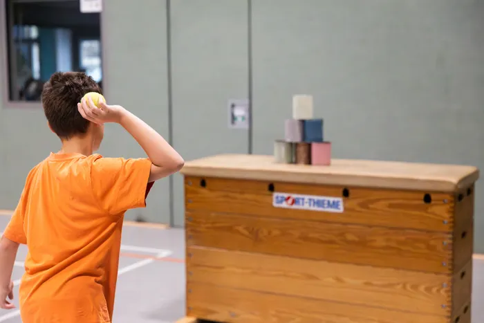 Ein Junge in einem orangefarbenen T-Shirt hält einen gelben Ball hinter dem Kopf und zielt auf einen Stapel kleiner Blöcke auf einer Holzkiste.