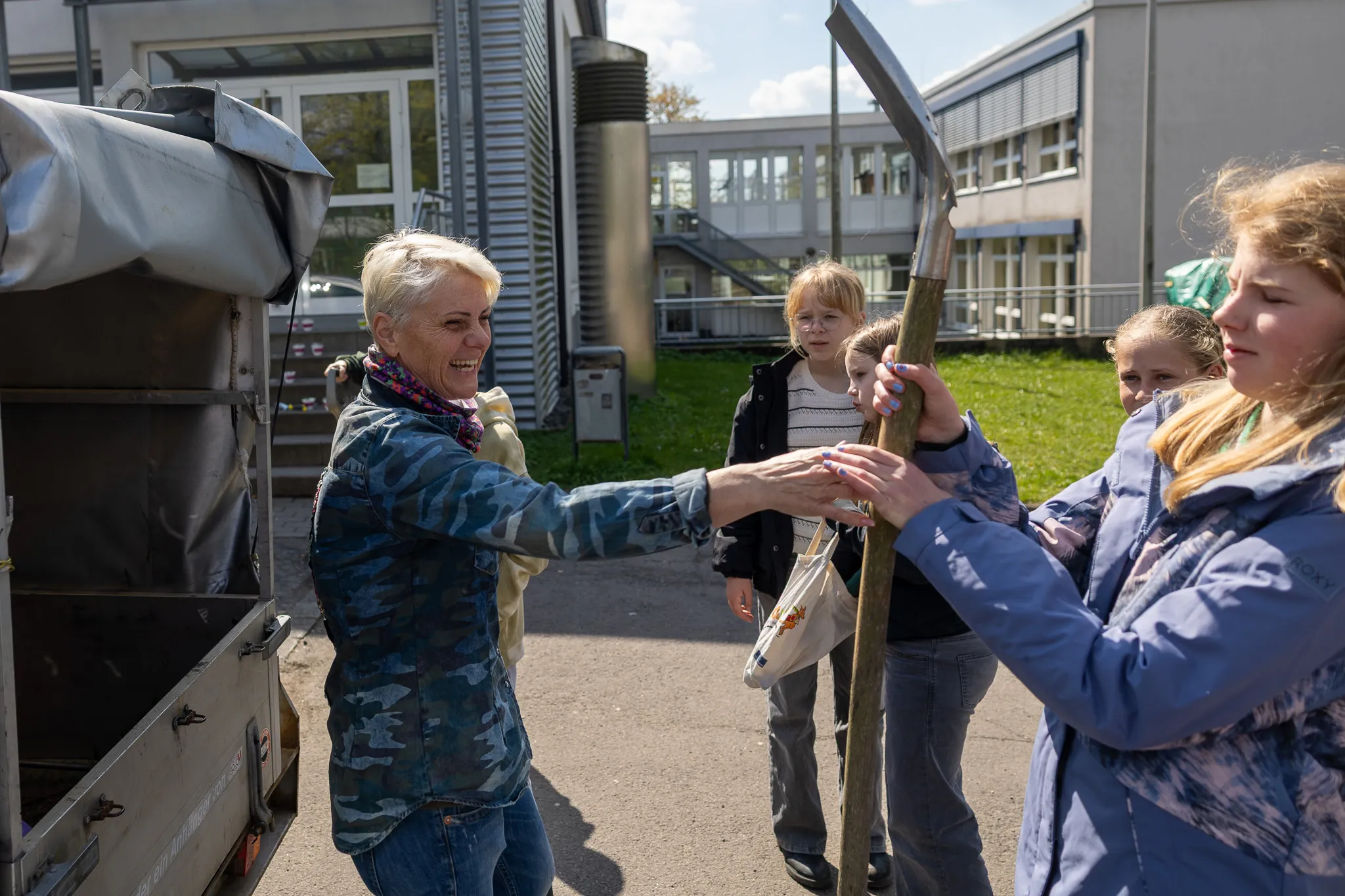 Eine Frau reicht einem Mädchen einen langen Stock, während andere Kinder im Hintergrund zusehen. Im Hintergrund ist ein Schulgebäude zu erkennen.