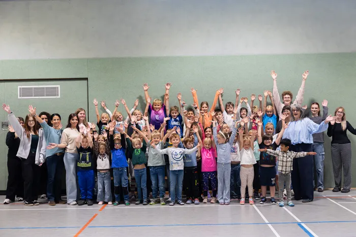 Eine große Gruppe von Kindern und Erwachsenen winkt in einer Turnhalle.