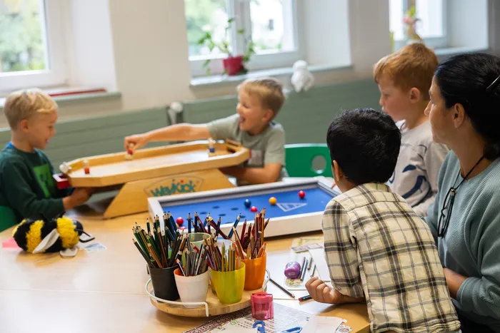 Kinder spielen an einem Tisch mit verschiedenen Gesellschaftsspielen, während eine Frau zuschaut.
