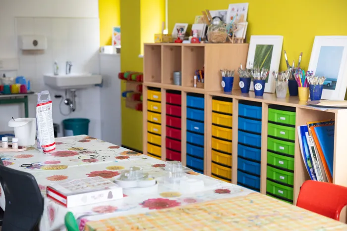 Ein Basteltisch mit Bastelmaterialien vor bunten Regalen und Schubladen mit Malutensilien.