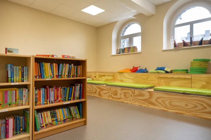 Ein Lesebereich mit Holzregalen voller Kinderbücher und einer langen Sitzbank mit grünen Polstern und bunten Kissen.