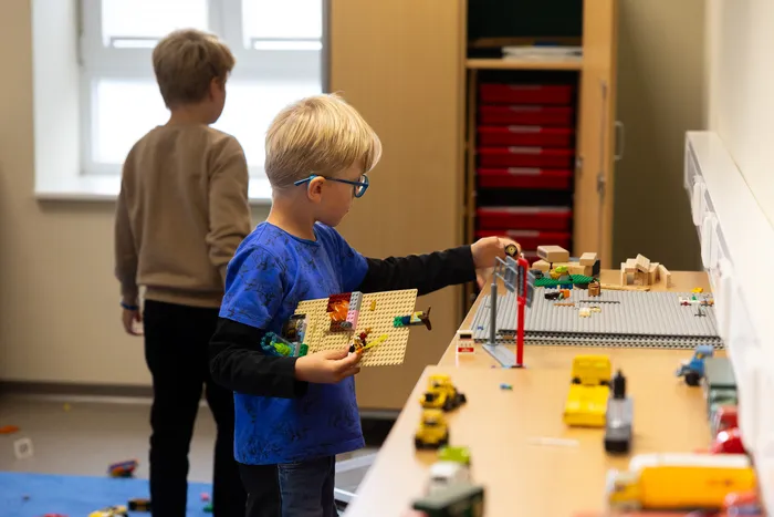 Ein Junge mit Brille spielt mit Bausteinen und Spielzeugautos auf einem Tisch.