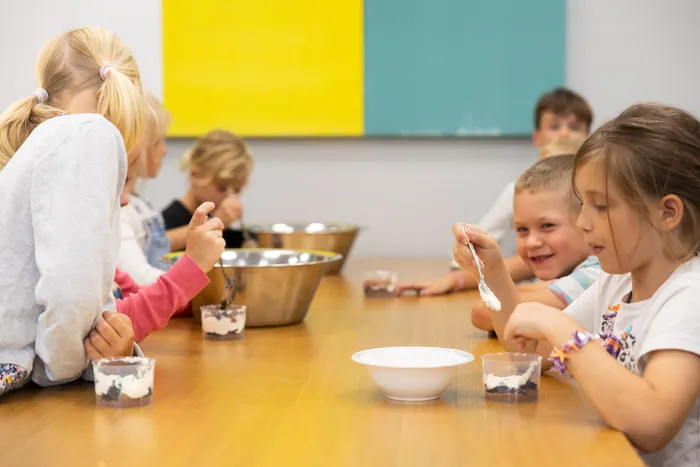 Kinder sitzen an einem Tisch und essen aus kleinen Bechern mit weißer Creme; auf dem Tisch stehen große Metallschüsseln.