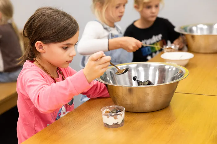Ein Mädchen schöpft mit einem Löffel Zutaten aus einer Metallschüssel in einen Becher, während im Hintergrund weitere Kinder Essen zubereiten.