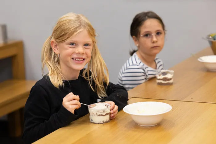Ein lächelndes blondes Mädchen hält einen Löffel und einen Becher mit Schichtdessert an einem Holztisch. Im Hintergrund sitzt ein weiteres Kind.