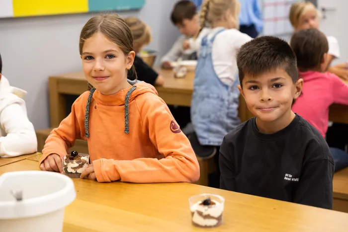 Ein Mädchen in einem orangefarbenen Hoodie und ein Junge in einem schwarzen Shirt sitzen an einem Tisch mit kleinen Dessertbechern in einem Klassenzimmer.