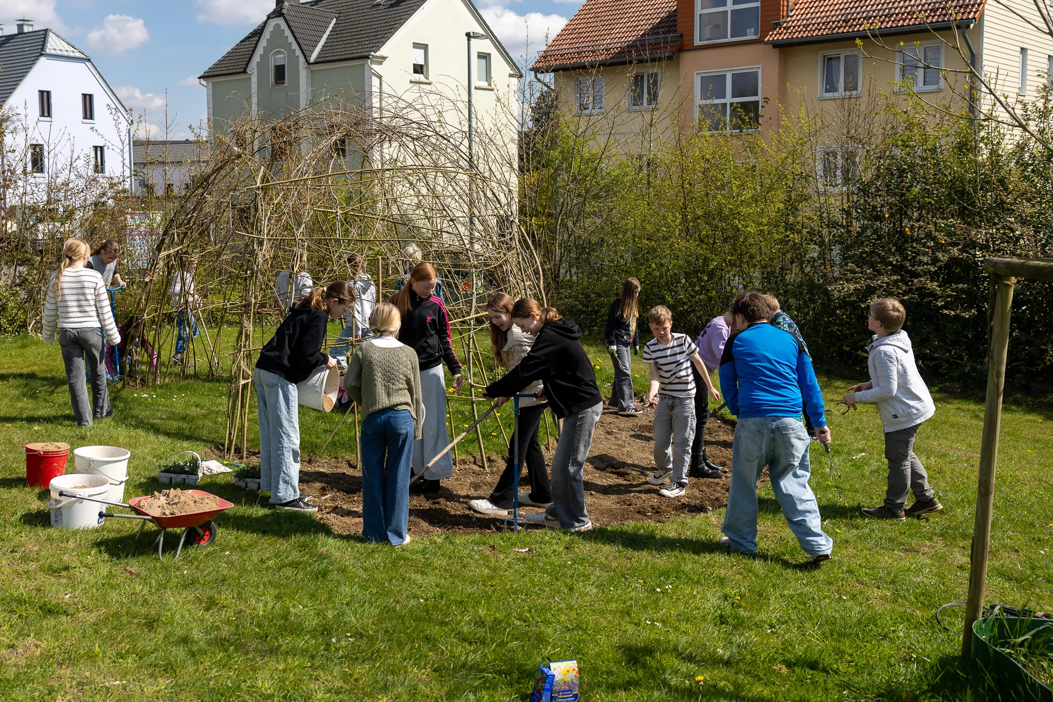Eine Gruppe Jugendlicher arbeitet in einem Garten, einige pflanzen einen Baum. Im Hintergrund sind Wohnhäuser und Büsche zu sehen.
