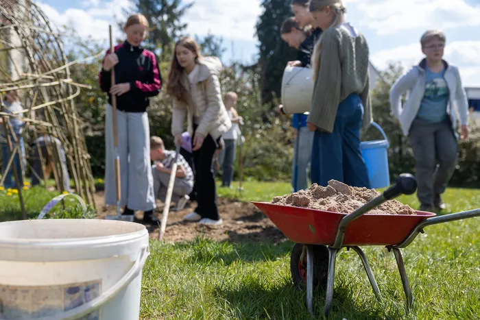 Eine Gruppe Jugendlicher arbeitet im Freien an einem Gartenprojekt, während eine Schubkarre mit Erde im Vordergrund steht.