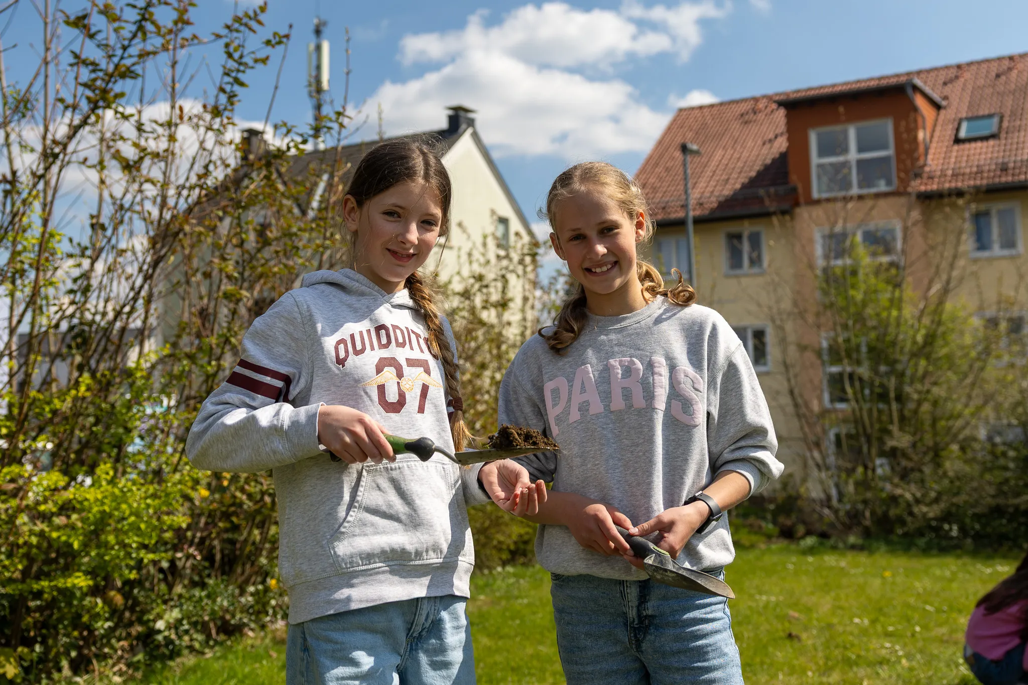 Zwei lächelnde Mädchen pflanzen im Garten vor einem roten Backsteingebäude. Sie tragen Hoodies mit Aufschriften und Jeans.