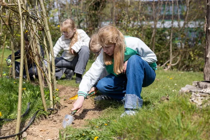 Zwei junge Frauen arbeiten im Garten. Eine kniet und pflanzt mit einem kleinen Spaten, die andere sitzt im Hintergrund und hält etwas in der Hand.