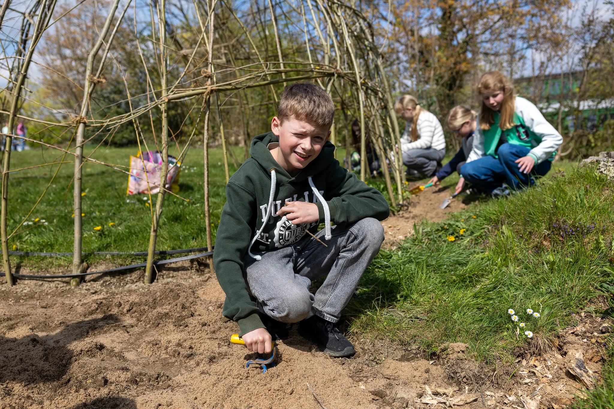 Ein Junge hockt im Gemüsegarten und pflanzt mit einer kleinen Schaufel. Im Hintergrund arbeiten weitere Kinder an den Beeten.