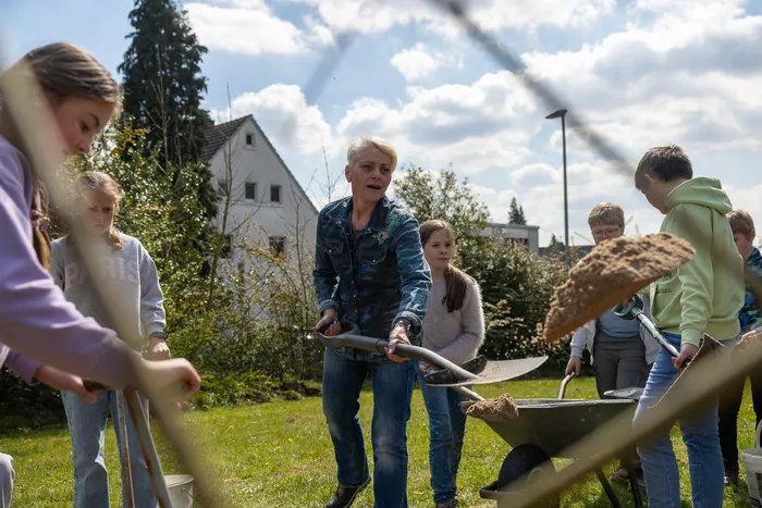 Eine Frau schiebt eine Schubkarre mit Erde, umgeben von Kindern bei einer Gartenaktion im Schulhof.
