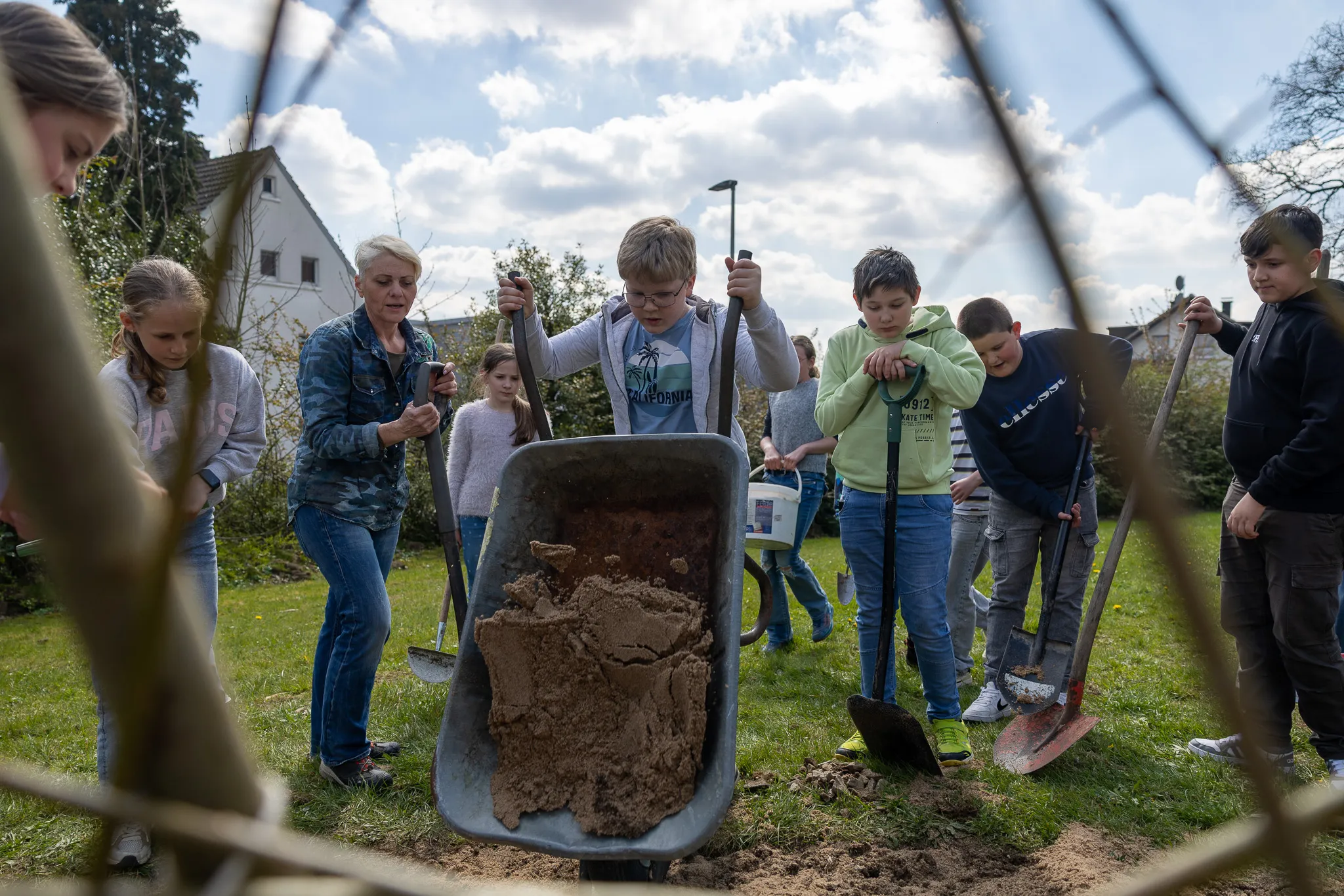 Eine Gruppe von Kindern und Erwachsenen arbeitet im Garten, ein Junge schiebt eine Schubkarre voller Erde. Im Hintergrund sind Häuser und ein Zaun zu sehen.