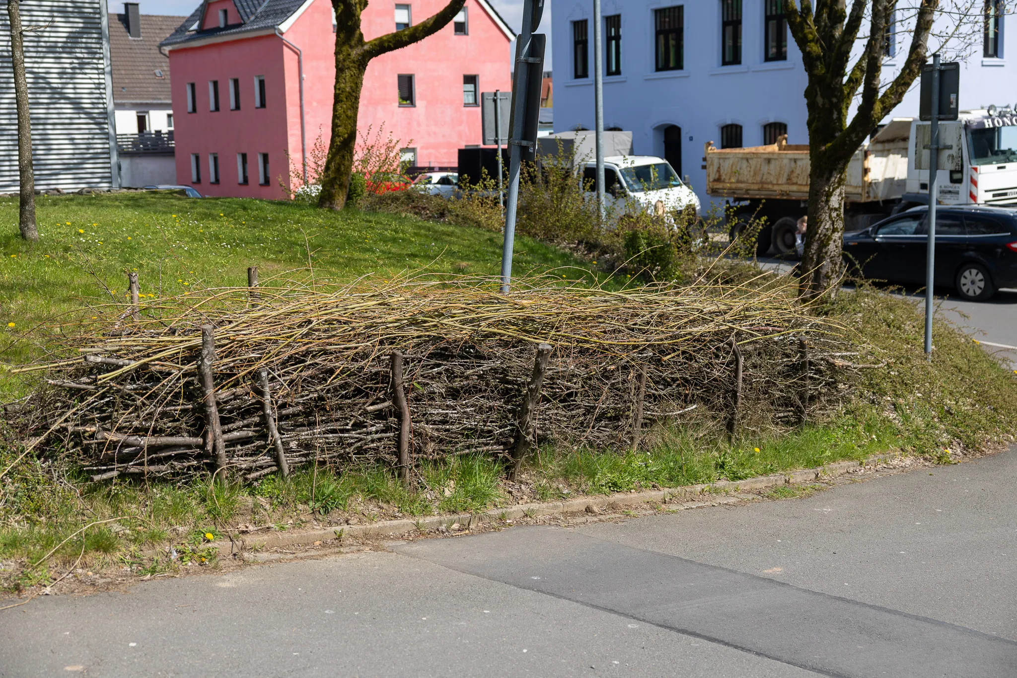 Ein provisorischer Zaun aus Ästen und Zweigen begrenzt eine Grünfläche neben einer Straße. Im Hintergrund sind Wohnhäuser in verschiedenen Farben und parkende Fahrzeuge zu sehen.