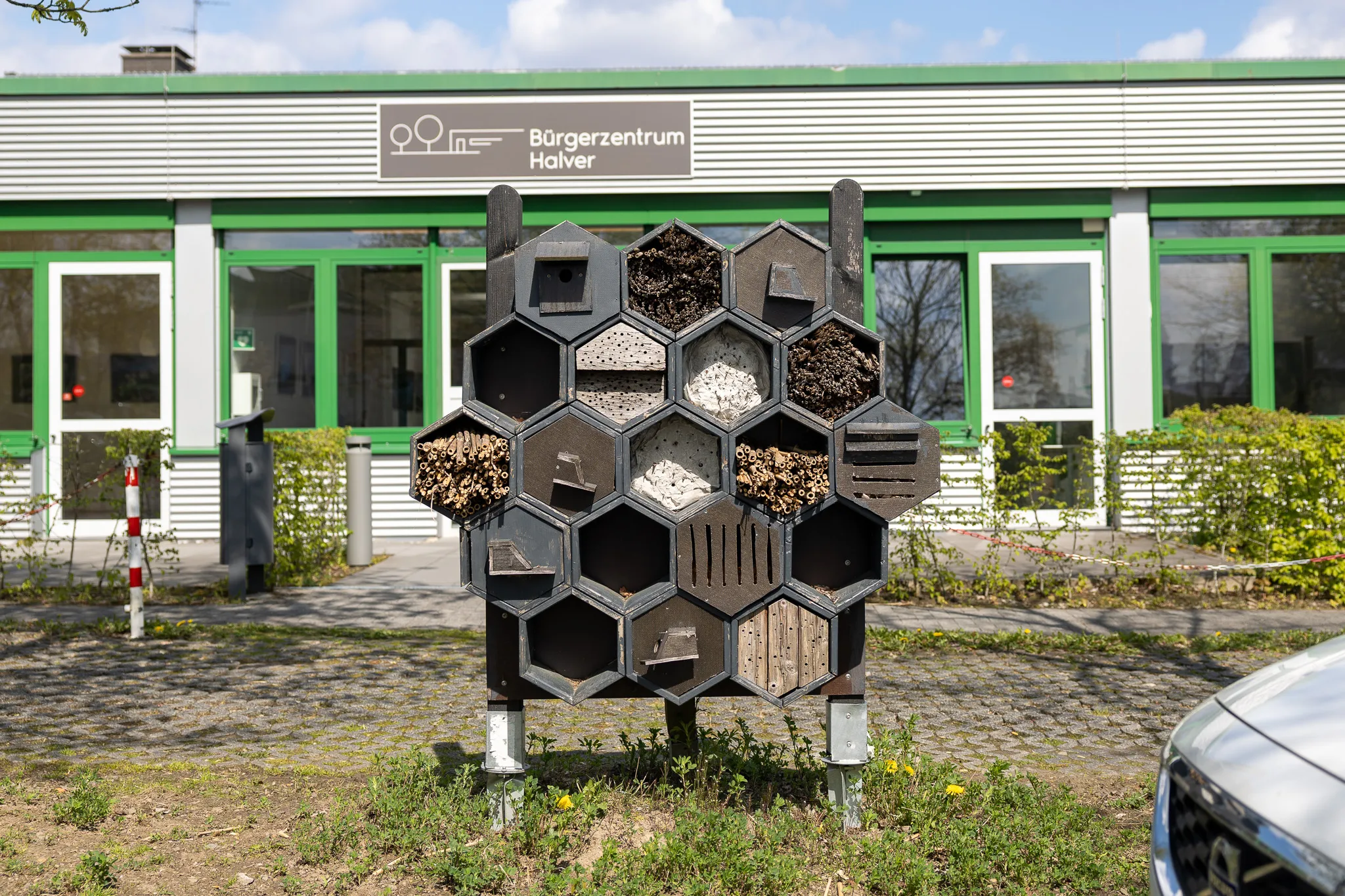Insektenhotel aus Holz vor dem Bürgerzentrum Halver, im Hintergrund ein Gebäude mit großen Fenstern und grünen Rahmen.