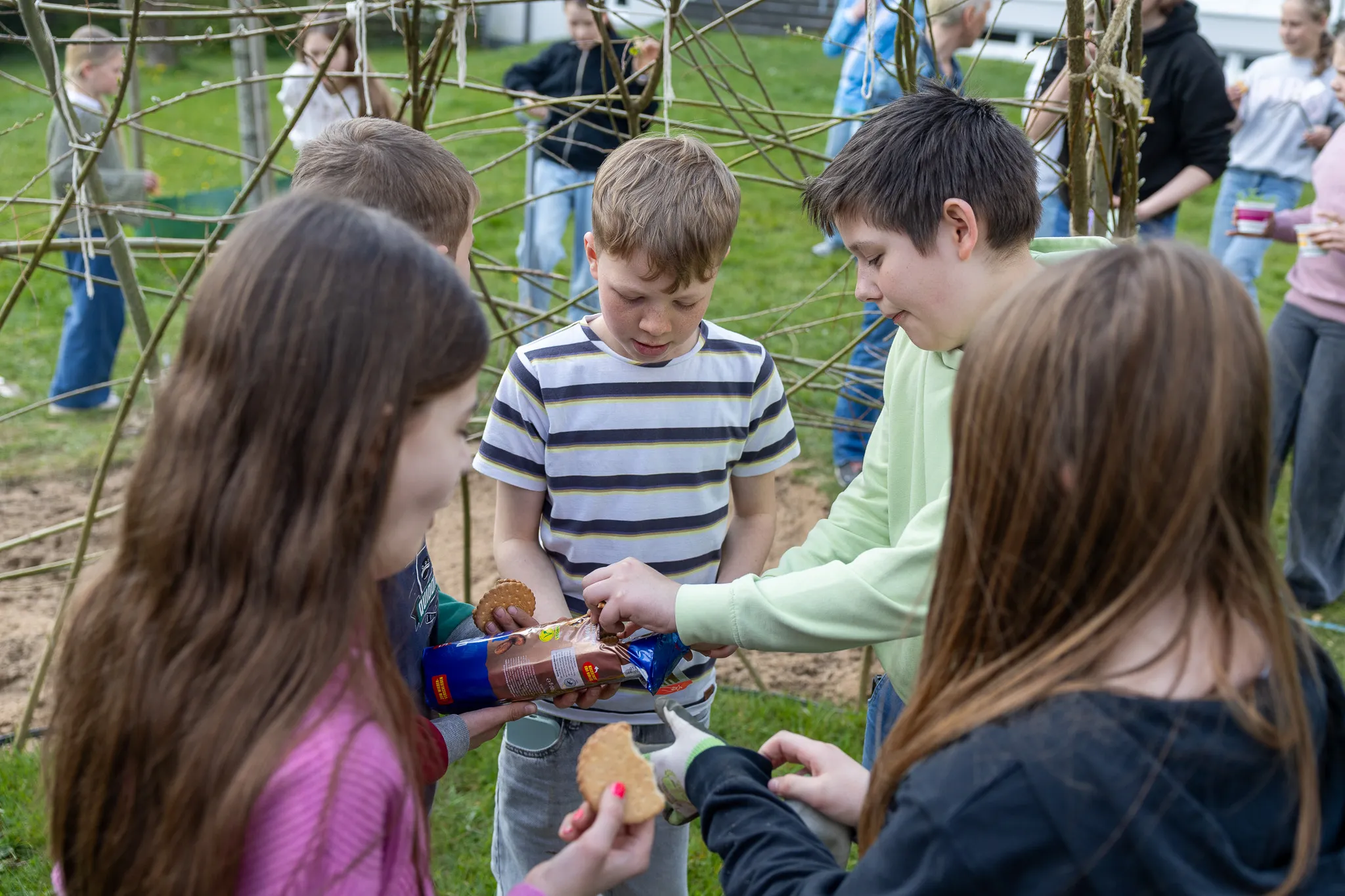 Mehrere Schülerinnen und Schüler stehen im Grünen und tauschen Kekse aus einer Packung aus.