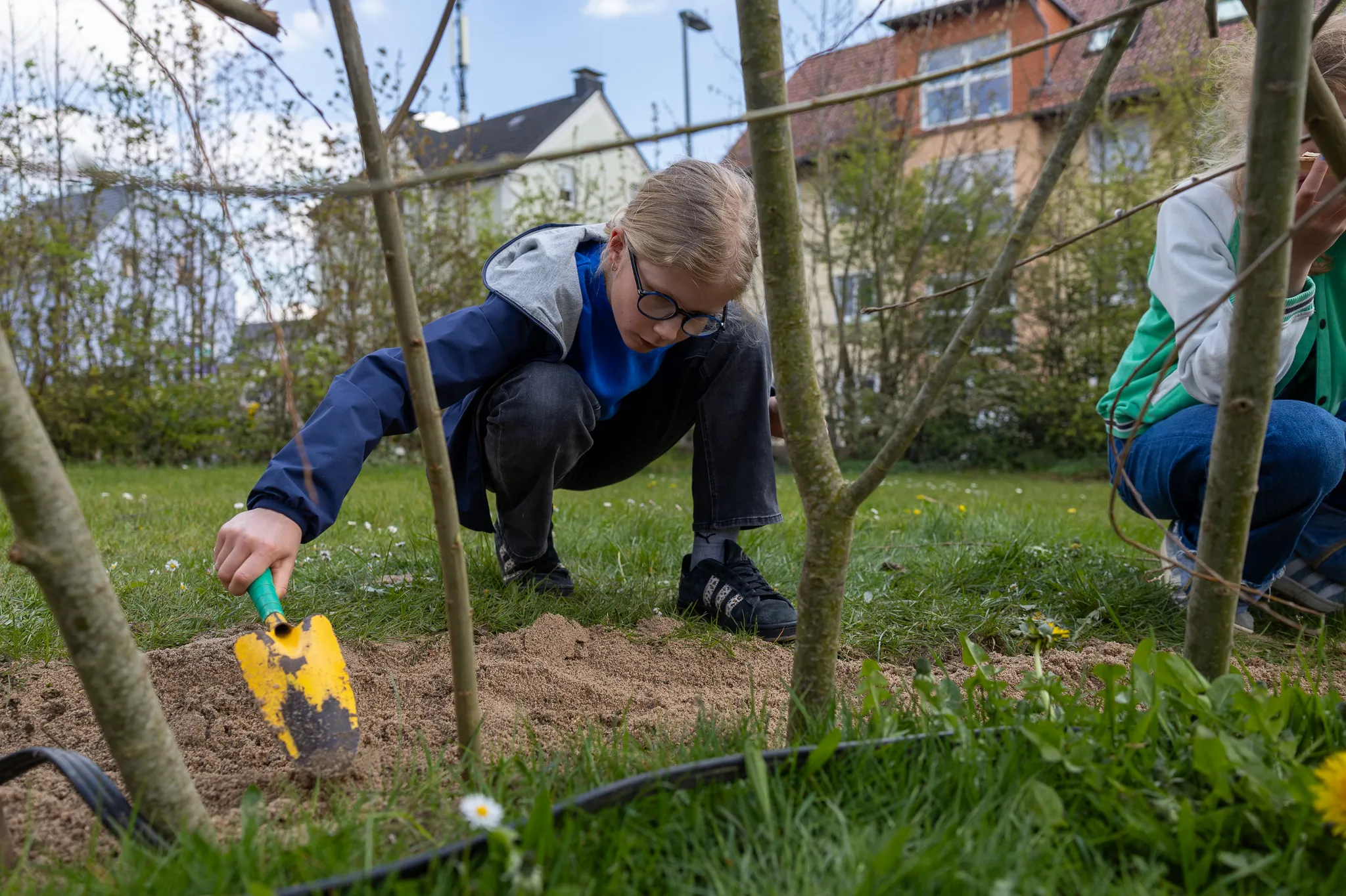 Ein Kind mit Brille kniet im Garten und gräbt mit einer kleinen Schaufel Erde um einen jungen Baum. Im Hintergrund ist ein weiteres Kind zu sehen.
