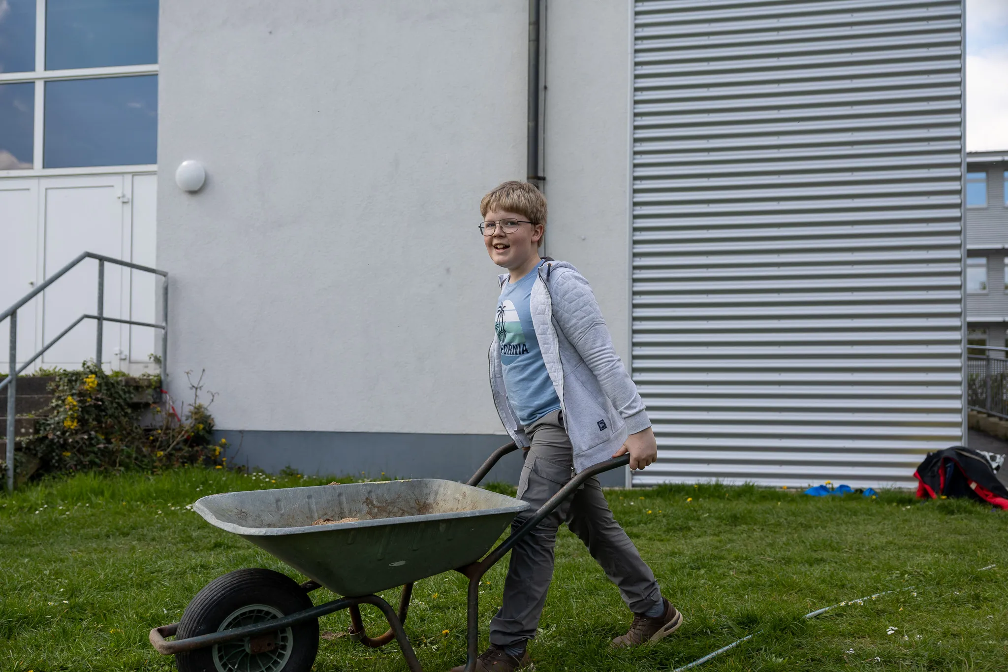 Ein Jugendlicher schiebt eine Schubkarre über einen Rasen vor einem hellen Gebäude mit großen Fenstern und einer Metallfassade.