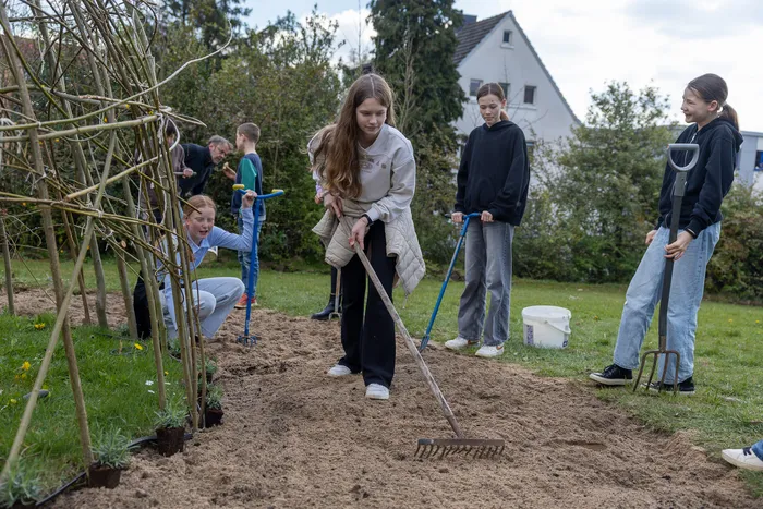 Mehrere Jugendliche arbeiten in einem Schulgarten, eine junge Frau sticht mit einer Grabegabel in die Erde. Im Hintergrund sind weitere Schüler und ein Rankgerüst zu sehen.